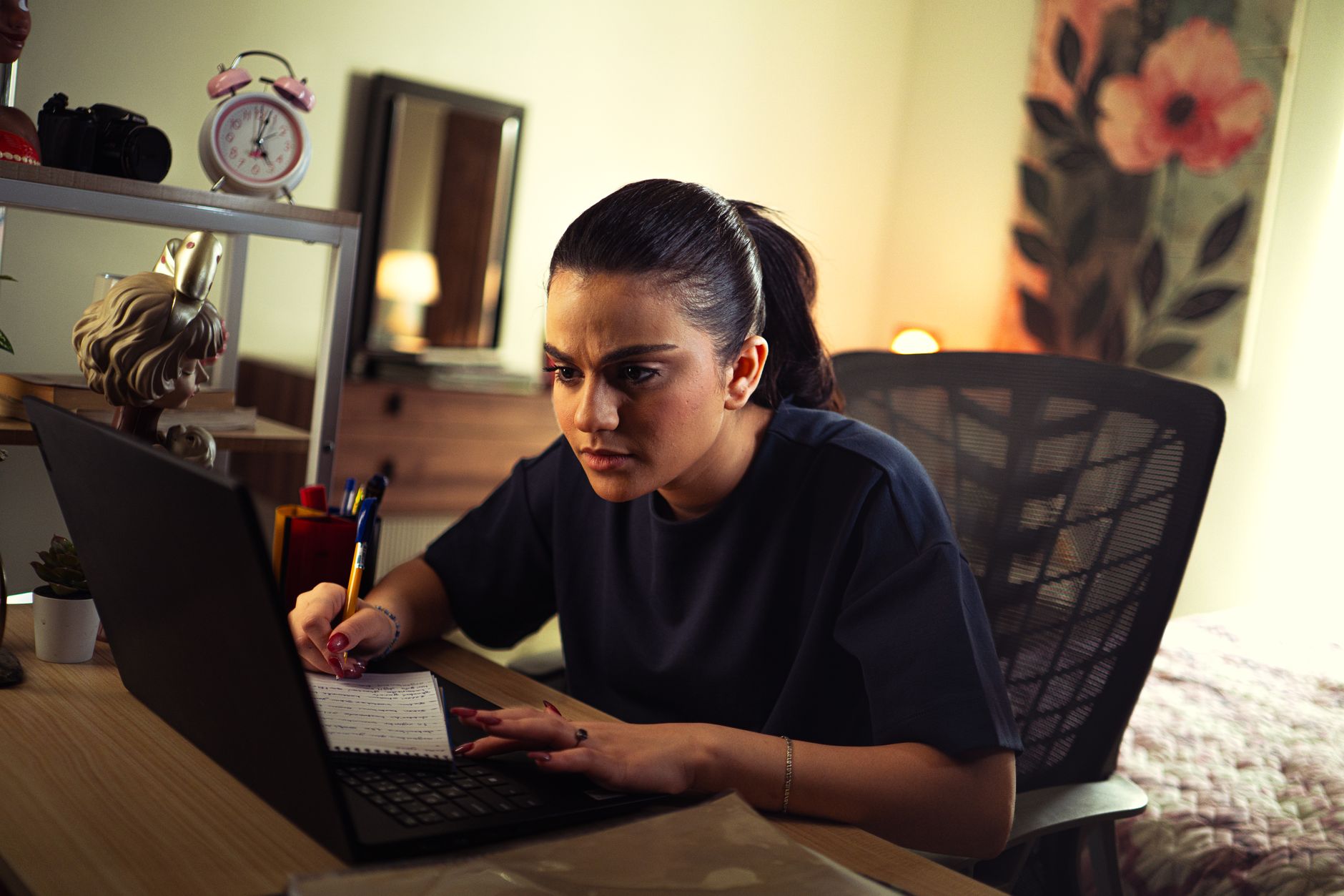 A young woman concentrating on a laptop at a desk in a warmly lit room at home, with a notepad and pen beside her.