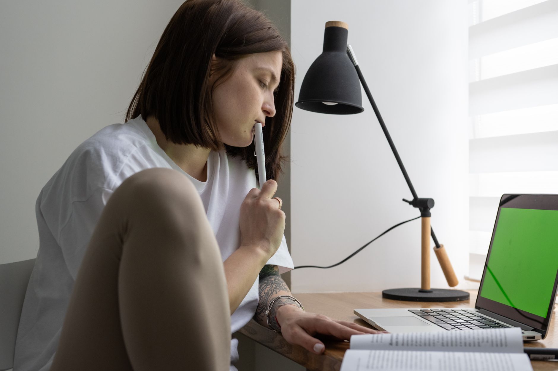 A thoughtful young woman seated sideways at a desk with a pen to her lips, a laptop and an open book in front of her in a bright room.