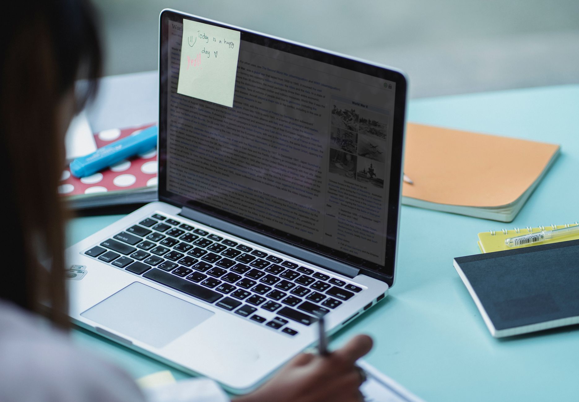 Over a woman's shoulder, an open laptop with a sticky note on the screen, beside notebooks and pens on a light blue desk.
